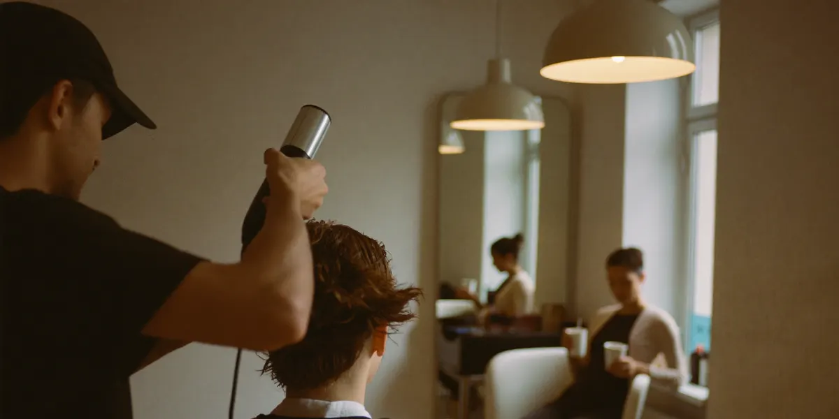 Une coiffeuse termine un brushing dans un petit salon, une cliente attend avec un café.